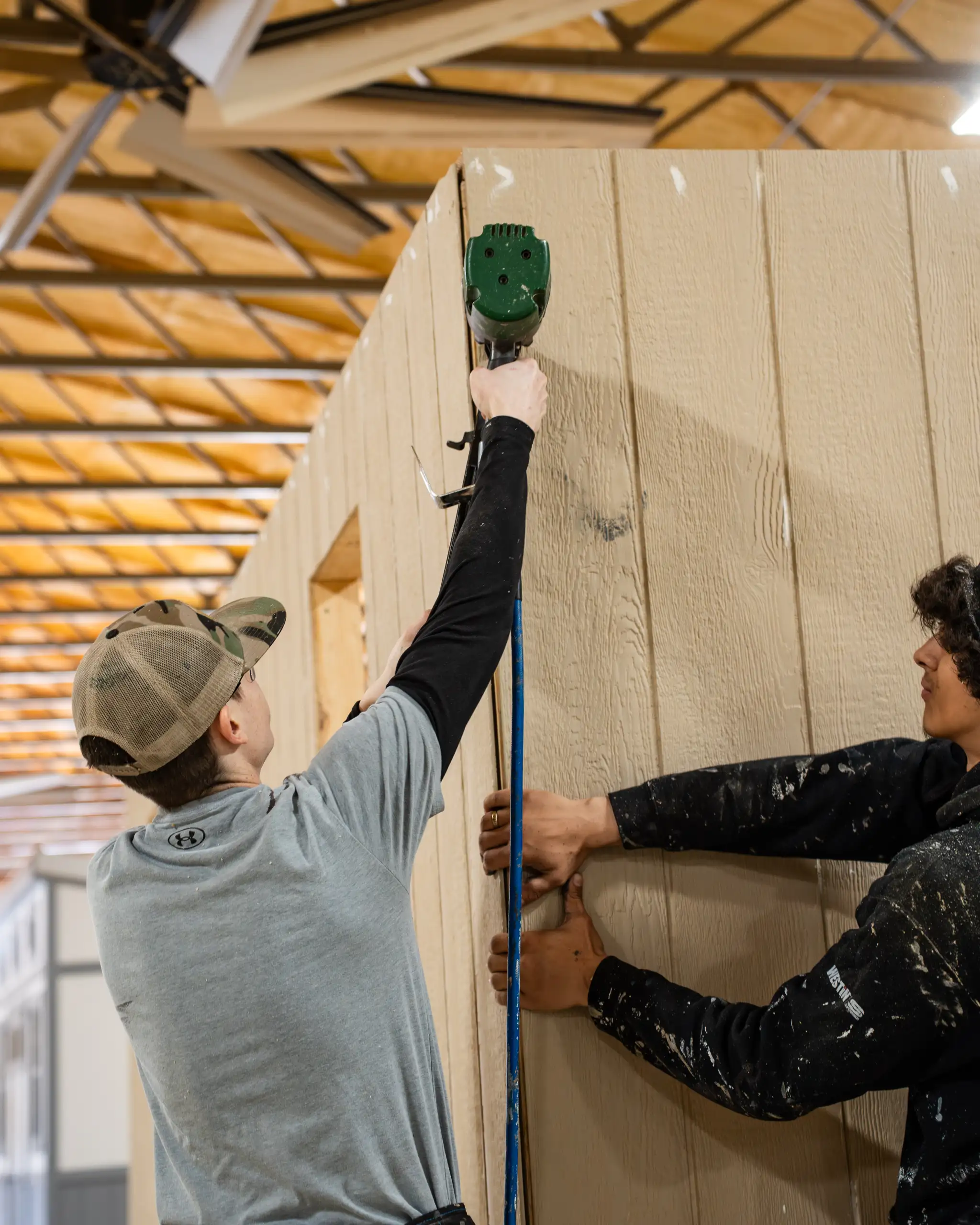 Two workers installing light-colored LP SmartSide panel siding on a shed wall using a nail gun inside a workshop