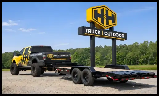 A yellow and black H&H Truck and Outdoor pickup truck towing a flatbed trailer in front of the company's large road sign.